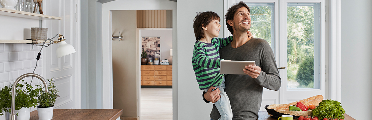 A father happily holding his son with a tablet in the other hand, looking around in a kitchen